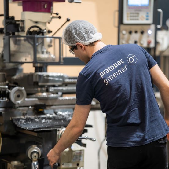 Ein Mann mit Haarnetz und Brille bedient eine Maschine in einer Fabrik. Die Maschine ist groß und mit Metallteilen gefüllt. Er trägt ein blaues T-Shirt mit dem Text 'pratopat gmein'.
