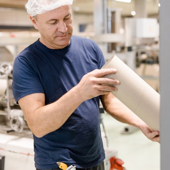 Ein Mann in einem blauen T-Shirt überprüft ein gerolltes Stück Papier in einem Fabrikgebäude. Er trägt eine Haarnetz.