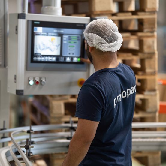 Ein Mann mit Haarnetz steht vor einer großen Maschine, den Rücken gekehrt, in einer Fabrik mit Metallstrukturen und Holzpaletten im Hintergrund.