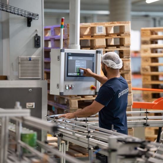 Ein Mann in einer Fabrik mit Haarnetz bedient ein Bedienfeld an einer Maschine. Im Hintergrund befinden sich Stapel von Holzpaletten.