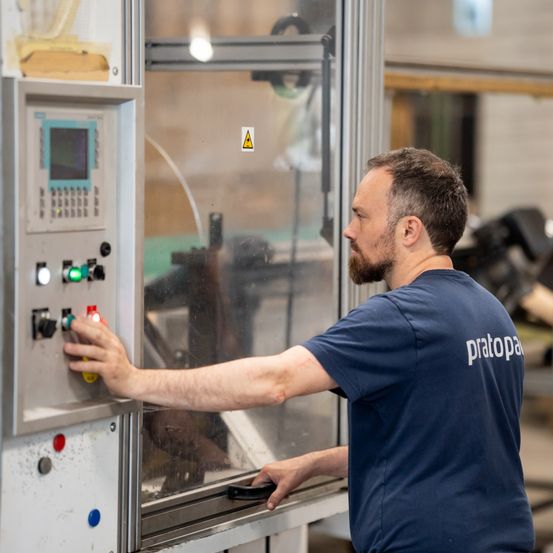 Ein Mann in einem blauen T-Shirt mit der Aufschrift 'pratopa' bedient eine Maschine mit verschiedenen Schaltern und Knöpfen in einer Fabrikumgebung.