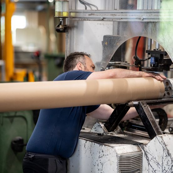 Ein Mann in einem blauen Shirt arbeitet an einer Maschine und schiebt einen großen zylindrischen Gegenstand durch eine Fabrik.