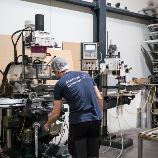 Ein Mann arbeitet an einer großen Maschine in einer Fabrik. Die Maschine hat ein Etikett mit der Aufschrift OPTIMUM. Er trägt eine Haarnetz und ein blaues Shirt mit den Worten pratopar gmeimer.