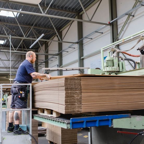 Ein Arbeiter steht auf einer Plattform in einer Fabrik und inspiziert gestapelte Kartonplatten. Im Hintergrund sind ein Förderband und Maschinen zu sehen.