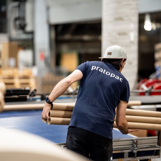 Eine Person mit einem 'pratopac'-Shirt arbeitet in einer Fabrik. Sie hält gerollte Papierröhren und trägt eine Mütze und eine Uhr.
