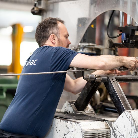 Ein Mann in einem blauen Shirt bedient eine große Maschine in einer Fabrik. Er hält eine Metallstange und scheint die Maschine einzustellen.