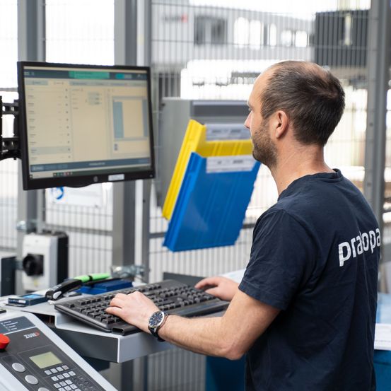 Ein Mann, der ein schwarzes Shirt mit dem Wort Prappa trägt, arbeitet in einer Fabrik an einem Computer. Er tippt auf einer schwarzen Tastatur und schaut auf den Monitor.