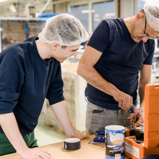 Zwei Männer in blauen Hemden und Haarnetzen arbeiten in einer geschlossenen Umgebung an einem Projekt. Ein Mann ist über einen Tisch gebeugt und der andere lehnt sich hinein, beide konzentrieren sich auf ein Objekt.
