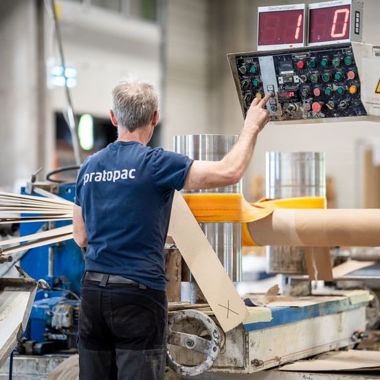 Ein Mann in einem blauen Shirt mit pratopac darauf bedient eine Maschine in einer Fabrik. Die Maschine hat zwei digitale Anzeigen mit Zahlen.
