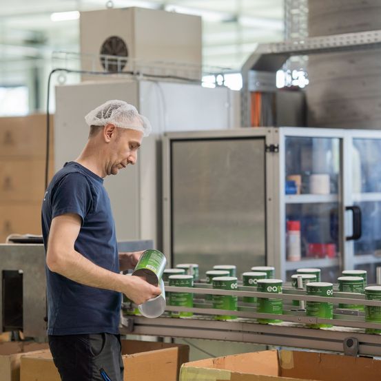 Ein Mann in einem blauen T-Shirt und Haarnetz handhabt eine grüne Dose auf einem Förderband in einer Fabrik. Hinter ihm befinden sich Maschinen und Lagerboxen.