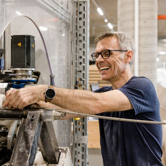 Ein Mann mit Brille und blauem T-Shirt arbeitet in einer Fabrik an einer Maschine. Er lächelt und scheint etwas zu reparieren. Die Maschine ist mit Kabeln verbunden und hat eine Metallstruktur.