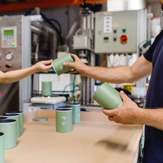 Zwei Personen in einem Fabrikambiente halten grüne Becher. Eine Person reicht einen Becher an die andere. Im Hintergrund sind Industriegeräte und ein Tisch mit mehreren grünen Bechern zu sehen.