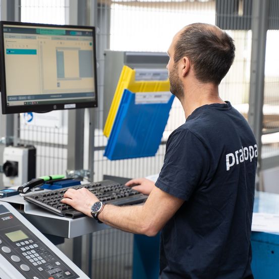 Ein Mann bedient einen Computer in einer Fabrikumgebung, trägt eine Uhr und ein blaues T-Shirt mit dem Wort 'praopa'. Der Monitor zeigt ein Diagramm.