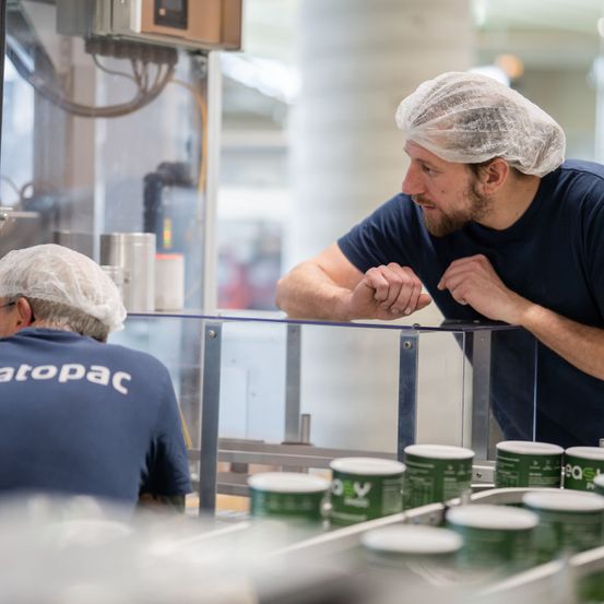 Zwei Männer in Haarnetzen und blauen Shirts, wahrscheinlich Fabrikarbeiter, beobachten ein Förderband mit grünen Behältern.