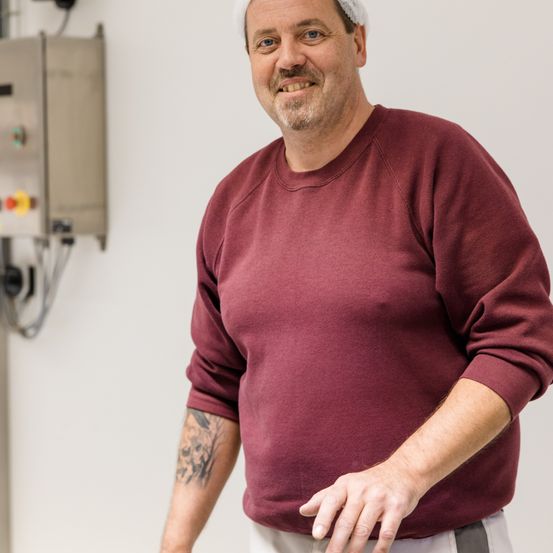Ein lächelnder Mann mit einem Tattoo am Arm steht in einem Raum mit weißen Wänden. Er trägt ein weinrotes Sweatshirt und weiße Hosen. Hinter ihm befindet sich ein graues Bedienfeld.