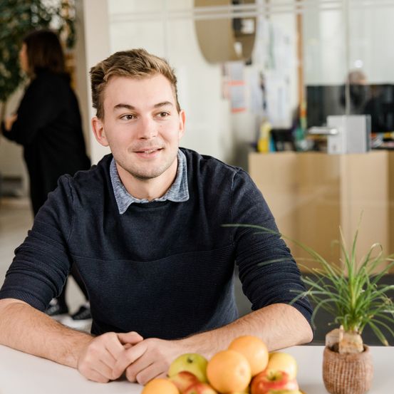 Ein junger Mann mit kurzen Haaren sitzt an einem Schreibtisch mit einer Schüssel Obst. Er lächelt und lehnt sich mit den Händen auf dem Tisch vor. Hinter ihm geht eine Frau.