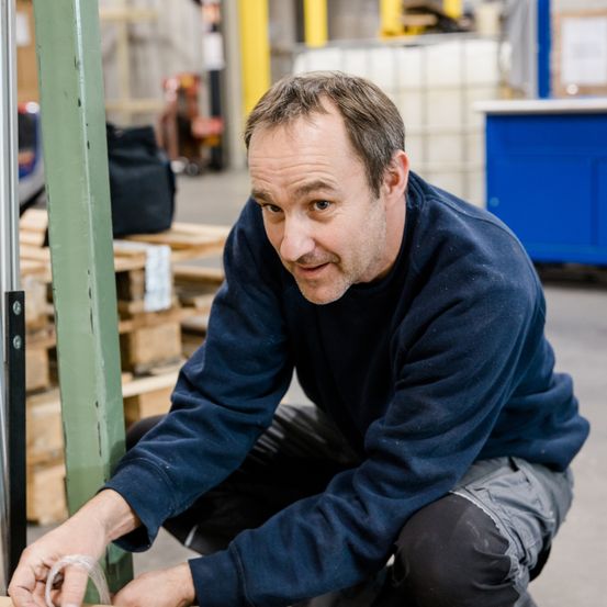 Ein Mann in einem blauen Sweatshirt hockt und scheint etwas an einer grünen Metallstruktur in einem Lager zu reparieren.
