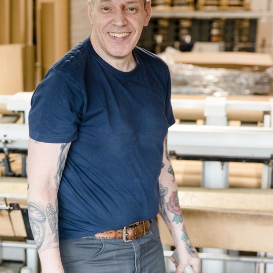 Ein Mann steht in einer Werkstatt und lächelt. Er trägt ein blaues T-Shirt, einen braunen Gürtel und hat sichtbare Tätowierungen. Hinter ihm sind Holzbänke und Lagerboxen.
