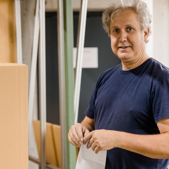 Ein Mann in einem blauen T-Shirt steht in einer Werkstatt und hält ein Blatt Papier in der Hand. Er sieht ernst aus. Hinter ihm sind Metallbalken und Holzbretter zu sehen.