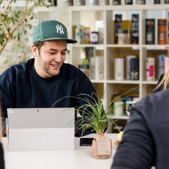 Ein lächelnder Mann in einem blauen Shirt und einer New York Yankees-Kappe sitzt an einem Schreibtisch mit Laptop, umgeben von Regalen voller Produkte.