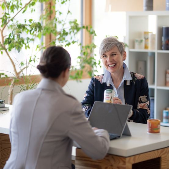 Zwei Frauen sitzen an einem Tisch, eine hält einen Laptop, die andere lächelt und hält eine Dose. Eine Pflanze und ein Regal sind im Hintergrund.