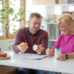 Ein Mann und eine Frau sitzen an einem Tisch, lächeln und halten Dosen und ein Papier in der Hand. Die Frau trägt ein rosa Shirt, der Mann ein Weinrot. Links stehen Äpfel auf einem Teller. Dahinter befindet sich ein Regal mit verschiedenen Gegenständen.