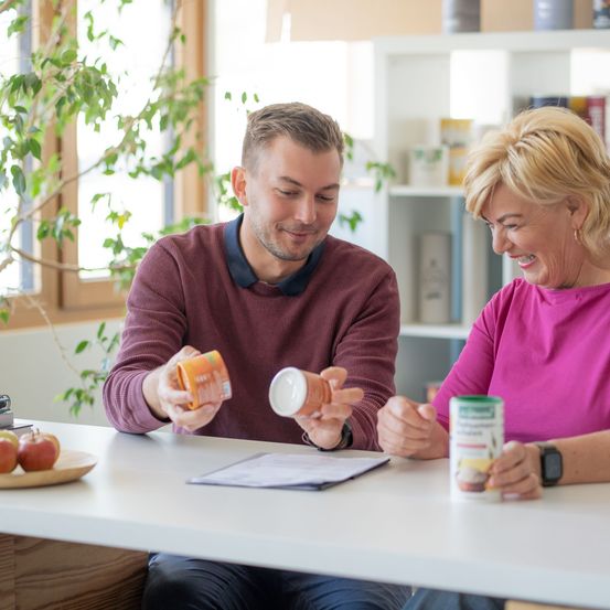 Ein Mann und eine Frau sitzen an einem Tisch, lächeln und halten Dosen und ein Papier in der Hand. Die Frau trägt ein rosa Shirt, der Mann ein Weinrot. Links stehen Äpfel auf einem Teller. Dahinter befindet sich ein Regal mit verschiedenen Gegenständen.
