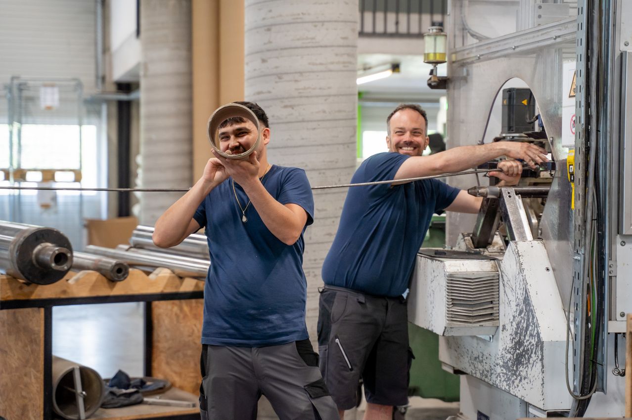 Two men in a workshop, one holding a round object and the other holding a long metal rod, both smiling and working on a machine.
