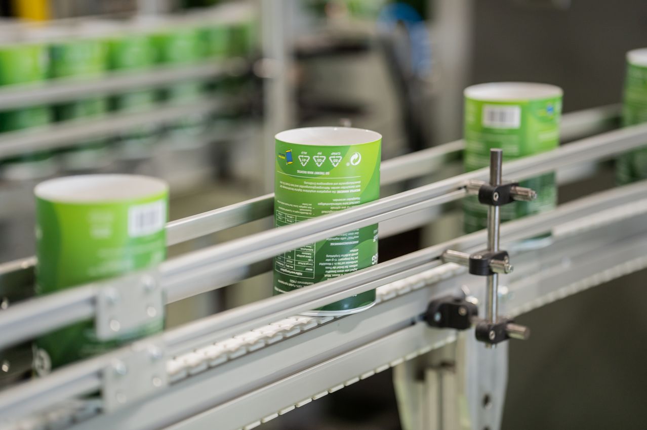 A green cylindrical container is on a conveyor belt in a factory. The container has a label with text and symbols.