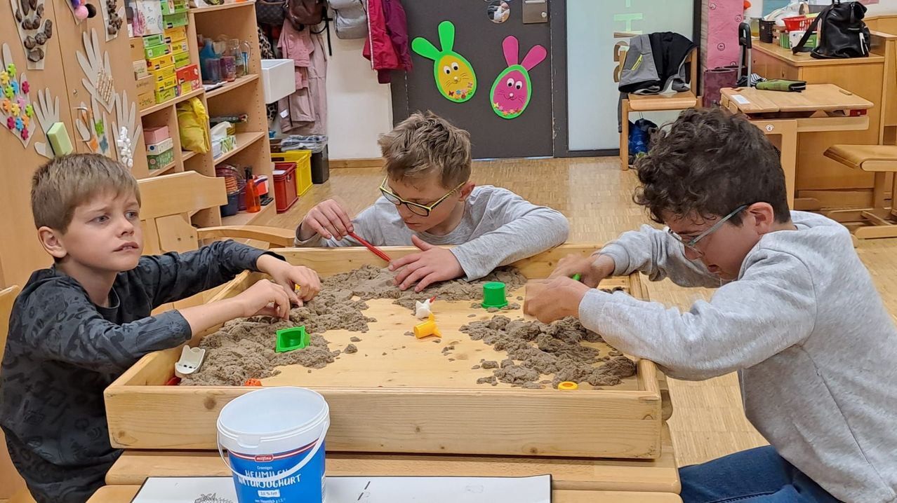 Drei junge Jungen spielen in einem Sandkasten mit kleinen Spielzeugen und Gegenständen. Der Junge in der Mitte trägt eine Brille und scheint sich auf die Aufgabe zu konzentrieren. Hinter ihnen befindet sich eine Wand mit Osterdekorationen und einem Aufbewahrungsbox.