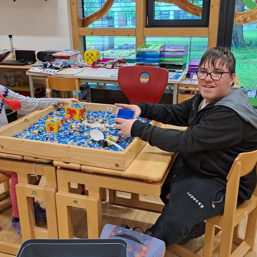 Ein Junge sitzt an einem Tisch und lächelt, während er mit einer Spielzeugkiste voller Plastikkugeln spielt. Neben ihm sitzt eine andere Person, möglicherweise ein Kind.