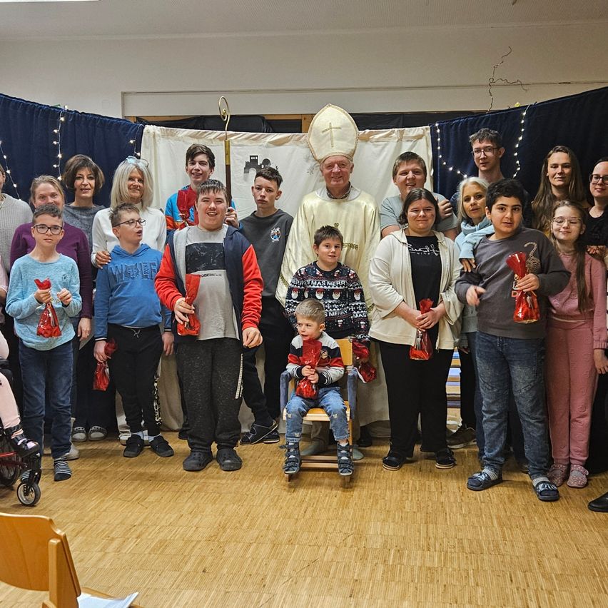Eine Gruppe von Menschen steht für ein Foto in einem Raum, mit einem Priester vorne. Kinder halten rote Taschen, und ein kleiner Junge sitzt auf einem Stuhl. Der Priester trägt eine weiße Robe. Blaue Vorhänge und Lichter sind im Hintergrund.