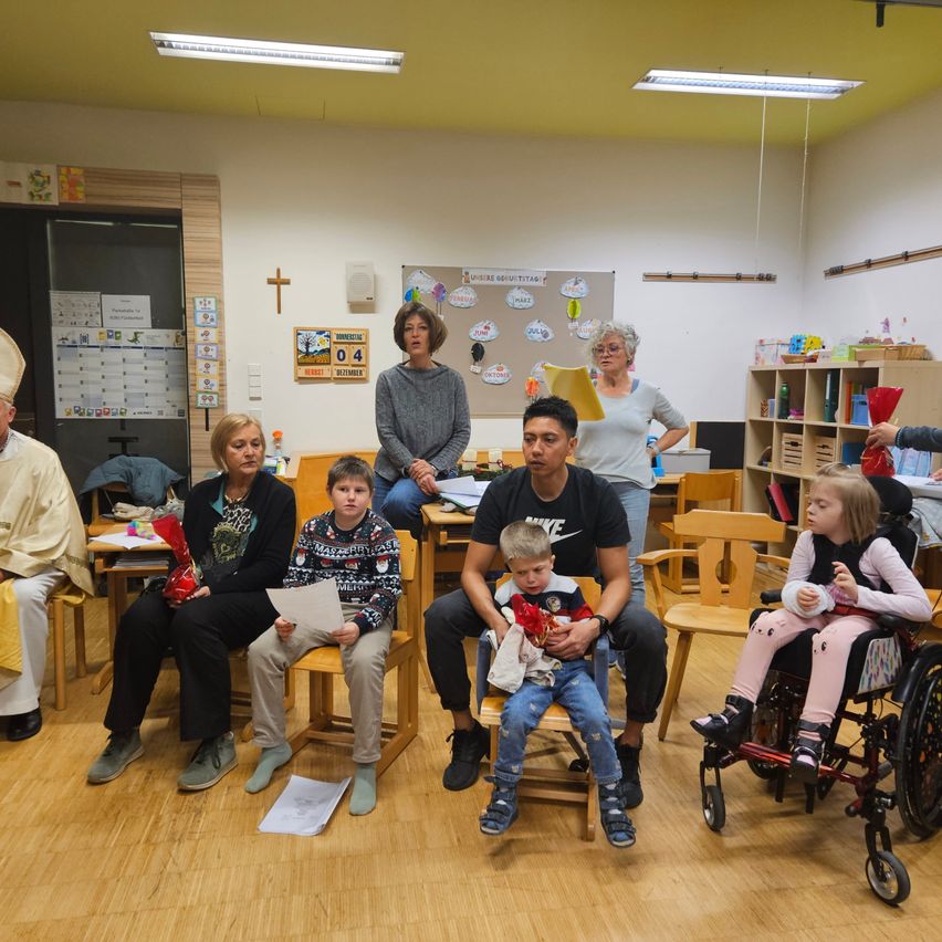 Eine Gruppe von Menschen sitzt in einem Klassenzimmer mit Holzböden. Ein Priester in weißen Gewändern ist anwesend, und das Klassenzimmer ist mit Postern und einer Bulletin-Board dekoriert. Einige Personen halten Papiere, während andere in Gespräche vertieft sind.