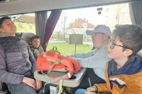 Vier Personen sitzen in einem Fahrzeug. Ein Mann in einer Jacke lächelt. Eine Frau mit Brille sitzt neben ihm. Ein Junge mit einem Rucksack schaut ihn an. Ein orangefarbener Rettungsring liegt auf dem Tisch.