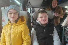 Zwei junge Personen sitzen in einem Bus. Das Mädchen trägt eine gelbe Jacke und eine graue Mütze, während der Junge einen schwarzen Weste und ein graues Langarmshirt trägt. Sie lächeln beide für die Kamera.