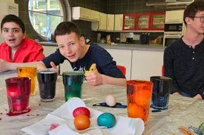 Drei Jungen sitzen in einer Küche um einen Tisch mit Gläsern und Ostereiern. Ein Junge lächelt, ein anderer zeigt, und der dritte sieht ernst aus. Bunte Flüssigkeiten und ein Ei liegen auf dem Tisch.