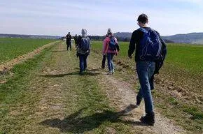 Eine Gruppe von Menschen mit Rucksäcken läuft auf einem Feldweg durch ein Feld bei klarem Himmel.
