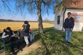 Drei Personen sind draußen, eine sitzt auf einer Bank, eine andere steht neben einem Baum und die dritte neben einem Haus mit einem roten Dach.