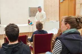 Ein Priester in einer weißen Robe steht vor einer Gruppe von Menschen, die in roten Stühlen in einem Raum sitzen.