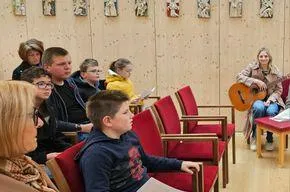 Eine Gruppe von Menschen, darunter Kinder und Erwachsene, sitzen auf roten Stühlen und hören aufmerksam einer Frau zu, die eine Gitarre auf einem Holzboden spielt.