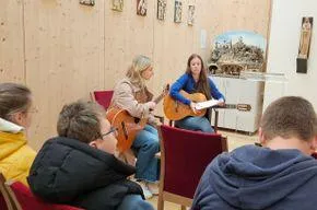 Eine Gruppe von Menschen sitzt in Stühlen in einem Raum. Eine Frau spielt Gitarre und eine andere hält ein Notenblatt. Hinter ihnen ist eine Holzwand und gerahmte Bilder.