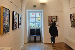 Eine Person geht durch eine Kunstgalerie mit Holzböden und weißen Wänden. Mehrere Gemälde sind an den Wänden ausgestellt. Zwei blaue Stühle stehen in der Mitte des Raumes.