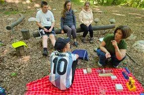 Eine Gruppe von Menschen sitzt auf einem Baumstamm und einer Decke im Wald. Eine Person trägt eine blaue Kappe und ein Trikot mit der Nummer 10. Eine Frau hält eine Flasche.