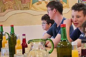 Mehrere Jungen sitzen an einem Tisch mit Getränken, darunter Orangensaft und eine Karaffe. Sie schauen sich etwas vor sich an. Hinter ihnen ist eine Wand mit einem Gemälde.