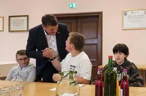 Ein Mann in einem Anzug schüttelt einem jungen Jungen in einem weißen T-Shirt die Hand. Sie sitzen an einem Tisch mit Gläsern, einer Karaffe und Flaschen. Hinter ihnen sitzt ein weiterer junger Junge und ein Mann in einer Jacke. Es gibt Bilderrahmen an der Wand.