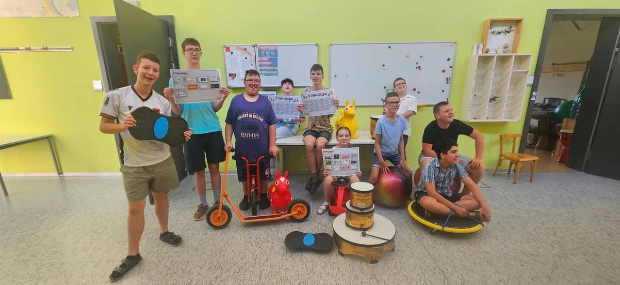 Eine Gruppe junger Menschen in einem Klassenzimmer, einige auf Übungsbällen sitzend, halten Urkunden und lächeln. Sie sind von verschiedenen Spielzeugen und Gegenständen umgeben.