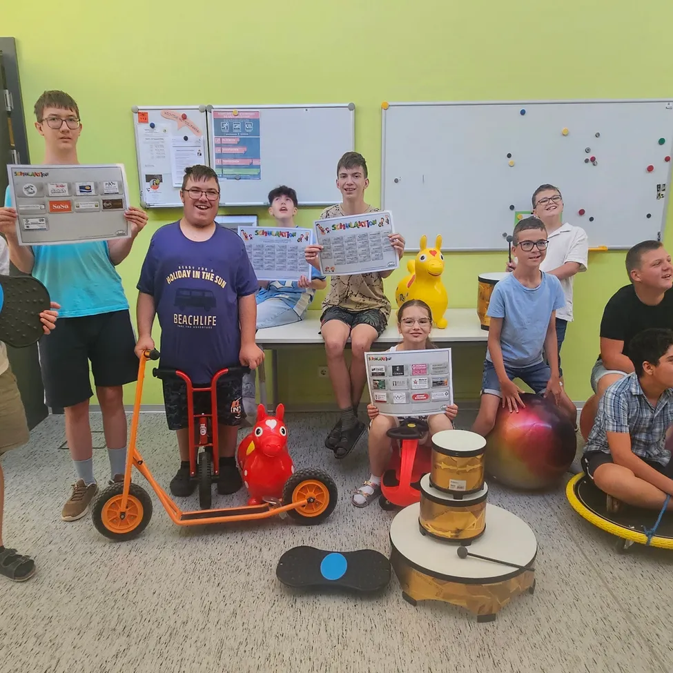 Eine Gruppe junger Menschen in einem Klassenzimmer, einige auf Übungsbällen sitzend, halten Urkunden und lächeln. Sie sind von verschiedenen Spielzeugen und Gegenständen umgeben.
