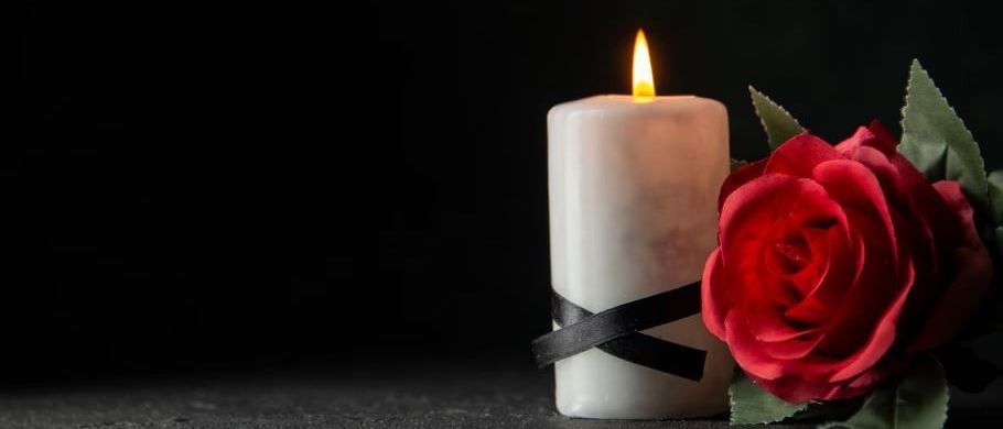 Eine brennende Kerze mit einem schwarzen Band neben einer roten Rose auf dunklem Hintergrund.