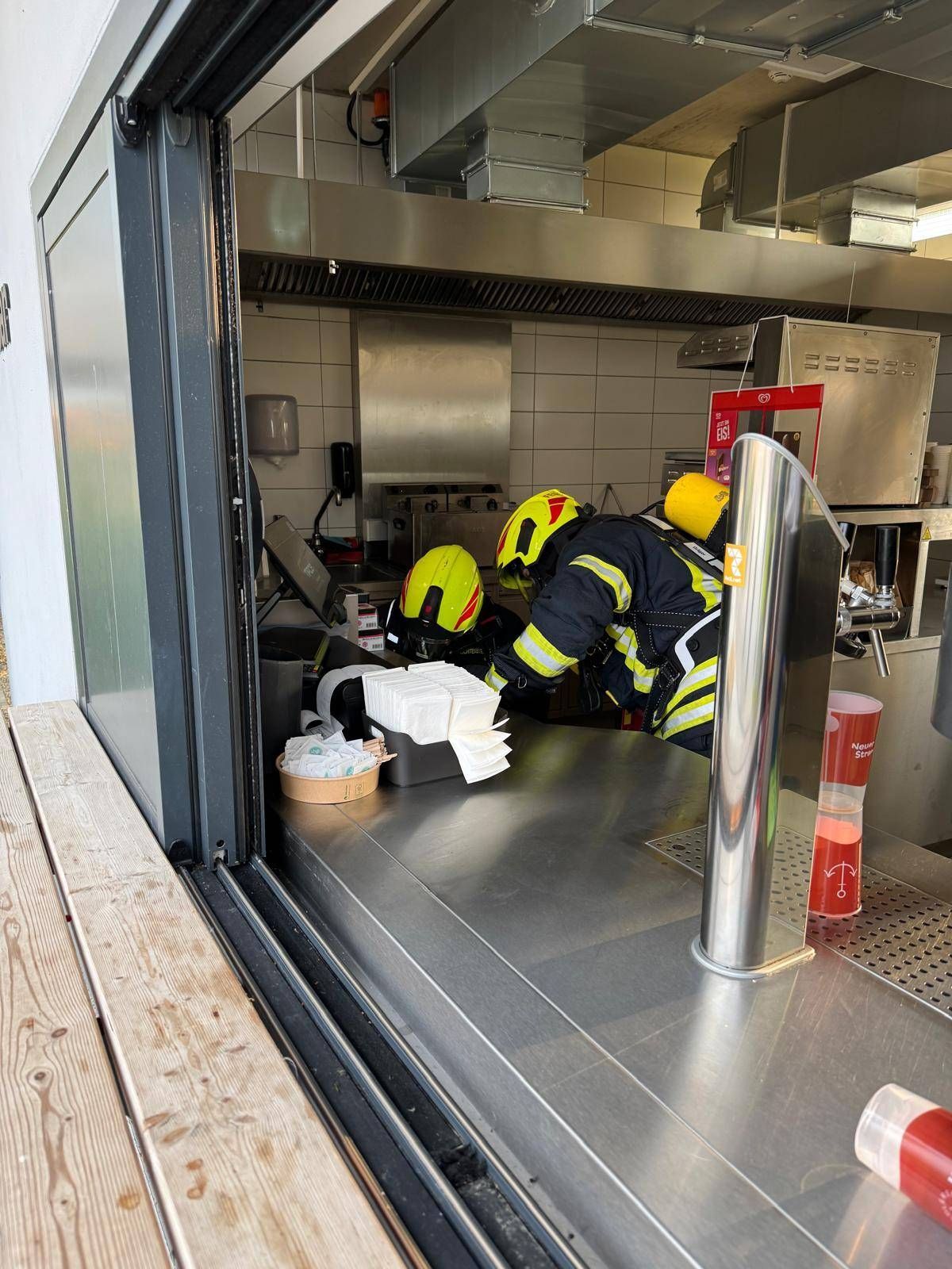 Zwei Feuerwehrleute in Schutzkleidung arbeiten in einer Küche, einer steht und der andere beugt sich vor. Es gibt mehrere Gegenstände auf der Arbeitsplatte.