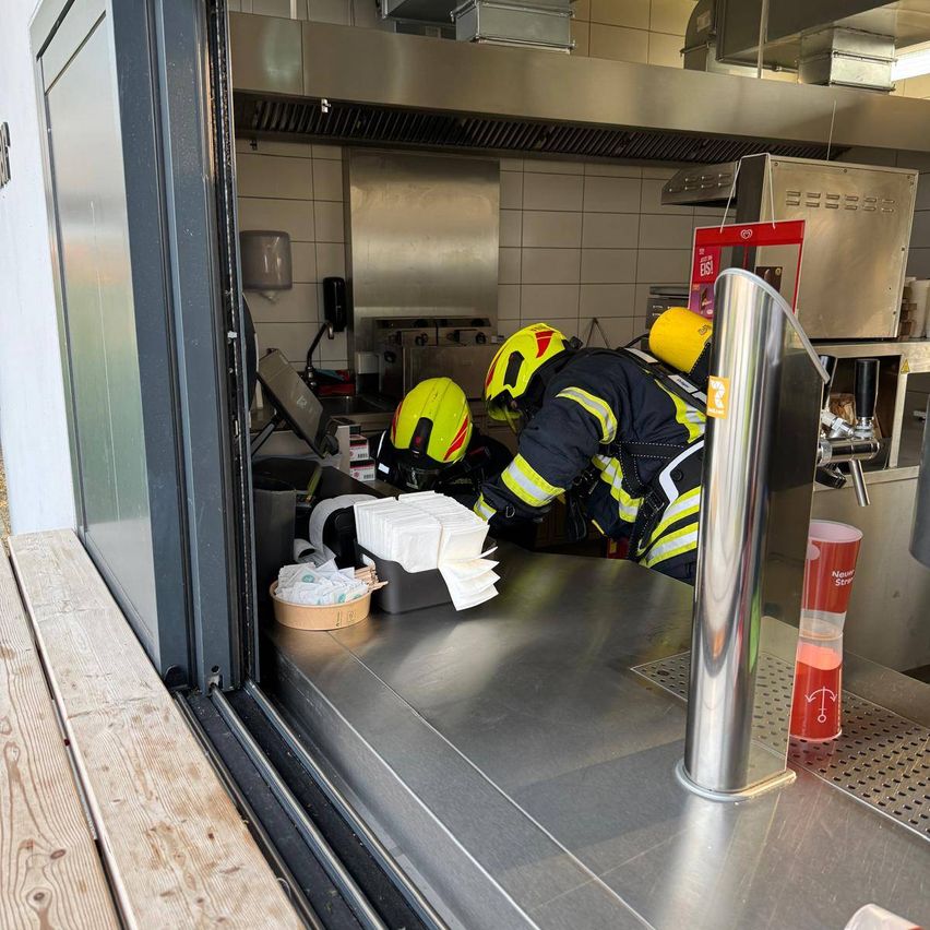 Zwei Feuerwehrleute in Schutzkleidung arbeiten in einer Küche, einer steht und der andere beugt sich vor. Es gibt mehrere Gegenstände auf der Arbeitsplatte.